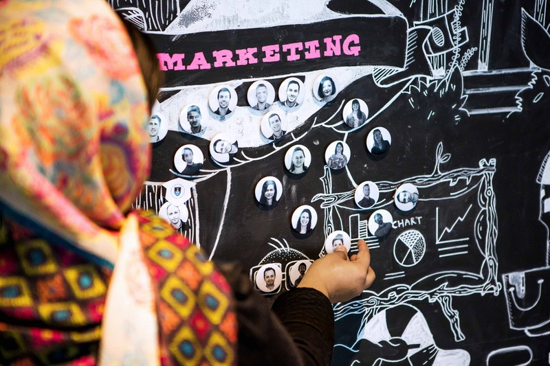 Woman wearing a head scarf adds a pin with a colleague's face on it to a mural with other pins on it titled 